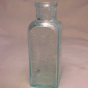 May include: A clear glass bottle with a textured surface. The bottle is rectangular and has a rounded top. The bottle is empty and has a light blue tint.