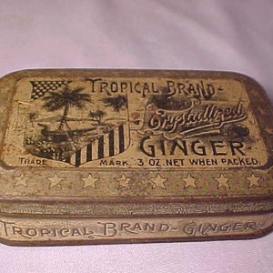 May include: A vintage tin box for Tropical Brand Crystallized Ginger. The box is made of metal and has a brown and gold color scheme. The front of the box features a design of a palm tree and a flag. The text "Tropical Brand Crystallized Ginger" is printed on the box in black lettering.