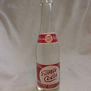 May include: A clear glass bottle with a red and white label that says "Tona Cola". The label also includes the text "Extra Carbonation" and "Caledonia Springs, Ltd."