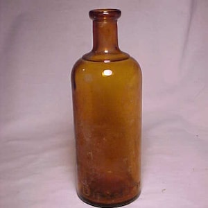 May include: A vintage amber glass bottle with a cylindrical body and a narrow neck. The bottle has a rounded shoulder and a dark brown hue. The glass appears slightly textured and shows signs of age.