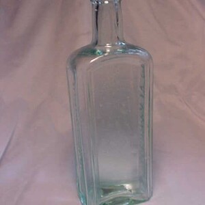 May include: A clear glass bottle with a square body and a rounded neck. The bottle is empty and has a slightly green tint.
