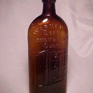 May include: A brown glass bottle with a label that reads "S.W.F. & Co. Kidney & Liver Cure". The bottle has a unique design with a raised image of a building.