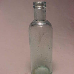 May include: A clear glass bottle with a rounded bottom and a slightly flared top. The bottle is empty and has a textured surface.