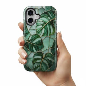May include: A smartphone case featuring a stained-glass design with large, green monstera leaves and gold-colored outlines. The case has a glossy finish and a protective bumper. The phone case is held in a hand against a white background.