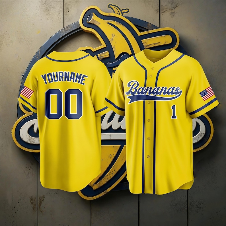 Op de afbeelding: Twee gele honkbalshirts met marineblauwe accenten. E&eacute;n shirt heeft "YOURNAME" en het nummer "00" op de rug. Het andere shirt zegt "Bananas" en heeft nummer "1". Beide shirts hebben een Amerikaanse vlag op de mouw.