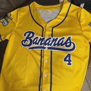 Op de afbeelding: Geel honkbalshirt met het woord "Bananas" in blauw schrift over de borst en het nummer 4 in blauw. Het shirt heeft blauwe randen en een teamlogo op de mouw.