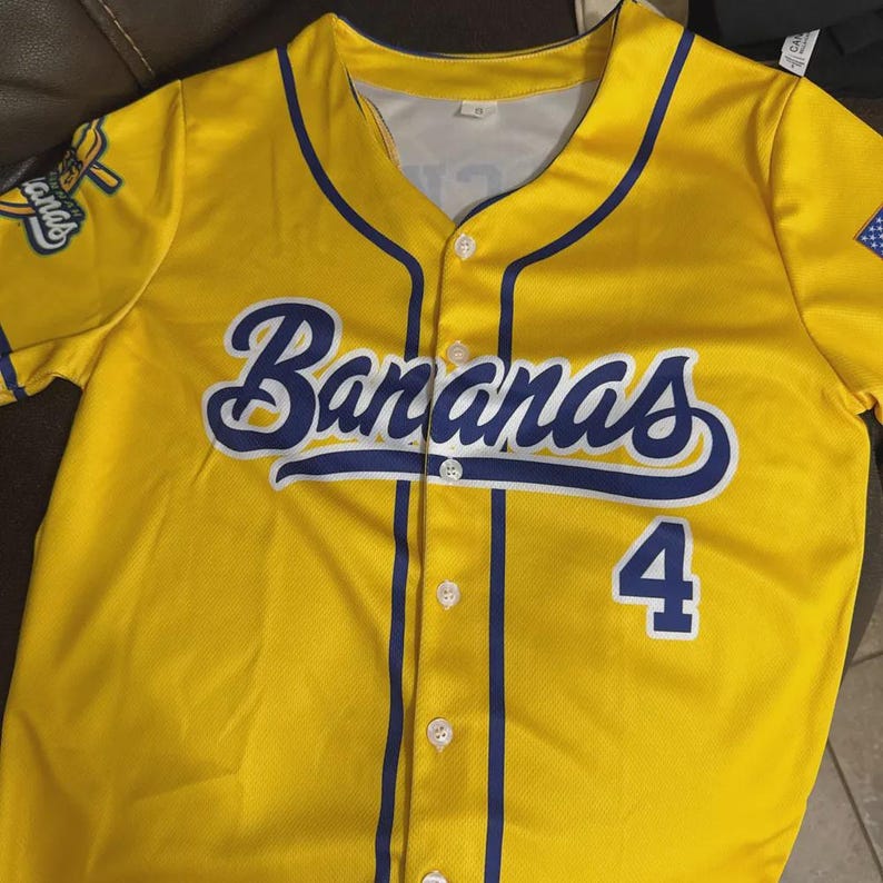Op de afbeelding: Geel honkbalshirt met het woord "Bananas" in blauw schrift over de borst en het nummer 4 in blauw. Het shirt heeft blauwe randen en een teamlogo op de mouw.