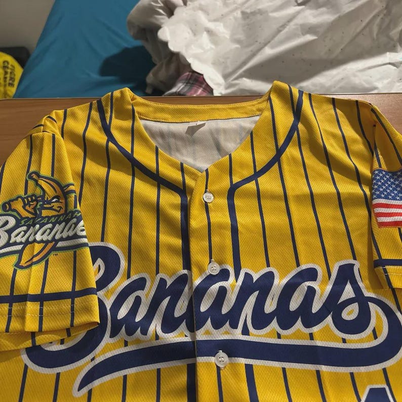 Op de afbeelding: Geel honkbalshirt met marineblauwe krijtstrepen en accenten. Het shirt heeft het woord "Bananas" in marineblauwe letters over de borst, samen met een teamlogo en een Amerikaanse vlag patch.