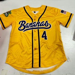 Op de afbeelding: Een geel honkbalshirt met het woord "Bananas" in marineblauwe letters over de borst en het nummer "4" eronder. Het shirt heeft marineblauwe biezen, knoopsluitingen en een Amerikaanse vlag op de mouw.