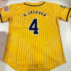 Op de afbeelding: Geel honkbalshirt met marineblauwe krijtstrepen, de naam "K. JACKSON" en het nummer "4" op de rug. Een Amerikaanse vlagpatch staat op de rechter mouw. Het shirt heeft een zwarte kraag en mouwboorden.
