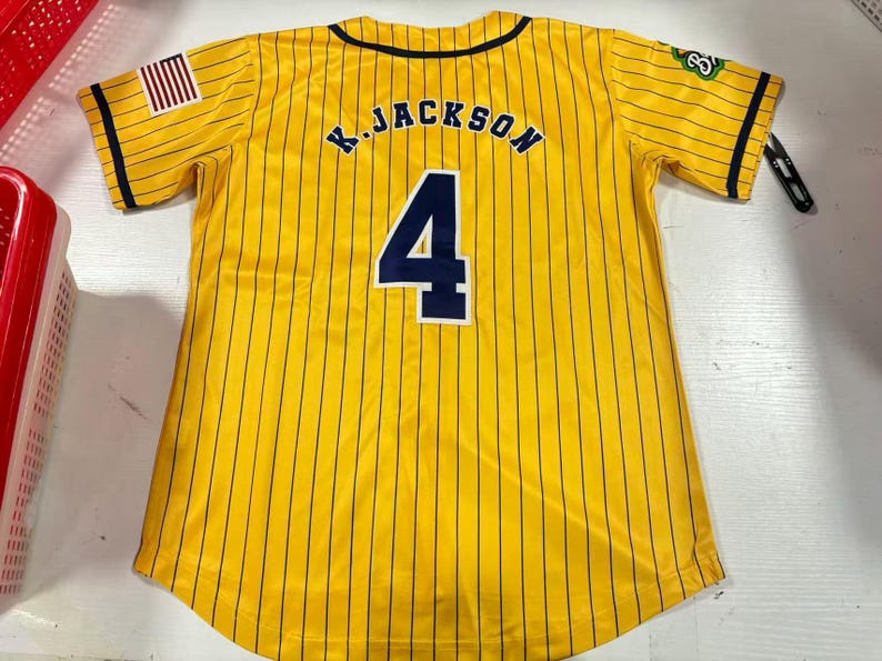 Op de afbeelding: Geel honkbalshirt met marineblauwe krijtstrepen, de naam "K. JACKSON" en het nummer "4" op de rug. Een Amerikaanse vlagpatch staat op de rechter mouw. Het shirt heeft een zwarte kraag en mouwboorden.