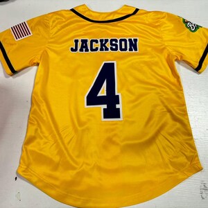 Op de afbeelding: Een geel honkbalshirt met de naam "JACKSON" op de rug en het nummer "4" in marineblauw. Het shirt heeft zwarte biezen en een Amerikaanse vlag op de mouw. Het shirt is gemaakt van een glanzend materiaal.