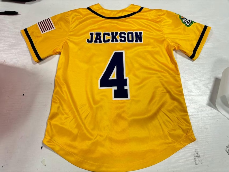 Op de afbeelding: Een geel honkbalshirt met de naam "JACKSON" op de rug en het nummer "4" in marineblauw. Het shirt heeft zwarte biezen en een Amerikaanse vlag op de mouw. Het shirt is gemaakt van een glanzend materiaal.