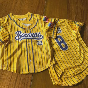 Op de afbeelding: Twee gele honkbalshirts met blauwe krijtstrepen. E&eacute;n shirt heeft het woord "Bananas" in blauw schrift en het nummer "23". Het andere shirt heeft het nummer "8". Beide shirts hebben kleine Amerikaanse vlaggen.