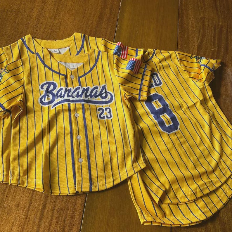 Op de afbeelding: Twee gele honkbalshirts met blauwe krijtstrepen. E&eacute;n shirt heeft het woord "Bananas" in blauw schrift en het nummer "23". Het andere shirt heeft het nummer "8". Beide shirts hebben kleine Amerikaanse vlaggen.