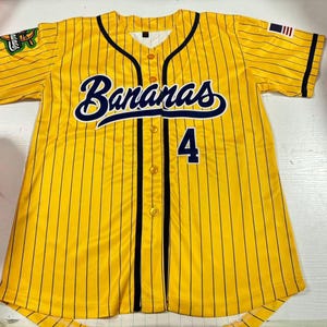 Op de afbeelding: Geel honkbalshirt met zwarte krijtstrepen, het woord "Bananas" in marineblauwe letters en het nummer 4. Voorzien van een kleine Amerikaanse vlagpatch en een teamlogo op de mouwen.