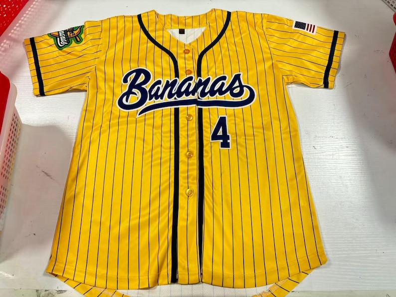 Op de afbeelding: Geel honkbalshirt met zwarte krijtstrepen, het woord "Bananas" in marineblauwe letters en het nummer 4. Voorzien van een kleine Amerikaanse vlagpatch en een teamlogo op de mouwen.