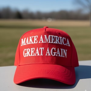 May include: A red baseball cap with the phrase "MAKE AMERICA GREAT AGAIN" embroidered in white. The cap is made of a smooth fabric and has a curved brim. The background is a blurred outdoor scene.