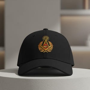 Può includere: Cappellino da baseball nero con un emblema ricamato raffigurante un sole, rami di alloro e un disegno a squadra e compasso in oro e rosso. Il cappellino è esposto su un piedistallo bianco.