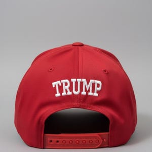 May include: Red baseball cap with the word "TRUMP" embroidered in white block letters. The cap has a red adjustable strap and a rounded top. The background is a solid gray.