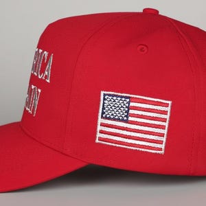 May include: A red baseball cap with the words "AMERICA AGAIN" embroidered in white. An American flag patch is on the side. The cap has a curved brim and a button on top.