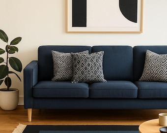 May include: A navy blue sofa with three decorative pillows featuring a geometric pattern. A framed abstract art piece hangs above the sofa. A rectangular rug with a blue gradient design is on the floor. A potted plant and a small wooden table are also in the room.