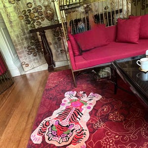 May include: A vibrant red rug with a tiger design in the center. The tiger features pink, red, and green accents. The rug is placed on a wooden floor, with a red floral patterned rug surrounding it. A red sofa and gold cage-like structure are in the background.