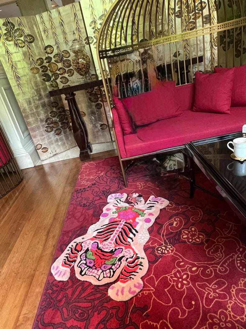 May include: A vibrant red rug with a tiger design in the center. The tiger features pink, red, and green accents. The rug is placed on a wooden floor, with a red floral patterned rug surrounding it. A red sofa and gold cage-like structure are in the background.
