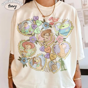 May include: Ivory Comfort Color t-shirt featuring a colorful graphic of Toy Story characters in heart-shaped frames. The design includes Buzz Lightyear, Woody, Jessie, and others, with floral accents and the words "Pizza Planet".