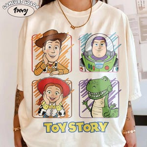 May include: Ivory Comfort Color t-shirt featuring four cartoon character portraits from Toy Story: Woody, Buzz Lightyear, Jessie, and Rex. The words "TOY STORY" are printed in blue below the characters. The shirt has a crew neck and short sleeves.