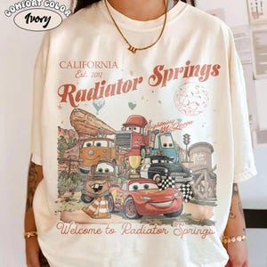 K&ouml;nnte beinhalten: Elfenbeinfarbenes T-Shirt mit einer Grafik mit Charakteren aus dem Film Cars. Das Design enth&auml;lt den Text "CALIFORNIA Est. 2012" und "Radiator Springs" im Vintage-Stil. Der Satz "Welcome to Radiator Springs" ist ebenfalls vorhanden.