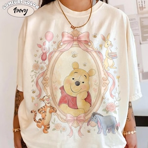 May include: Ivory-coloured t-shirt with a framed illustration of Winnie the Pooh and friends. The design includes Piglet with a balloon, Tigger, Eeyore, and Rabbit. The shirt has the words "Comfort Color" and "Ivory" printed on it.