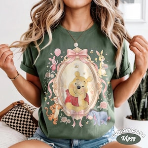 May include: Green t-shirt with a Winnie the Pooh design in an oval frame, alongside Piglet, Tigger, and Eeyore. The design includes pink bows, flowers, and the text "Comfort Color Moss".