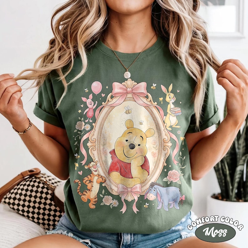 May include: Green t-shirt with a Winnie the Pooh design in an oval frame, alongside Piglet, Tigger, and Eeyore. The design includes pink bows, flowers, and the text "Comfort Color Moss".