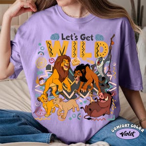 May include: A lilac-colored t-shirt featuring a graphic of characters from The Lion King. The design includes the text "Let's Get WILD" in yellow, along with illustrations of Simba, Mufasa, Scar, Timon, Pumbaa, and Zazu.