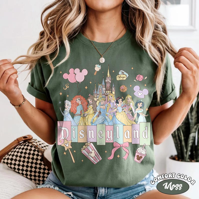 May include: Green t-shirt with a Disneyland graphic featuring Disney princesses, a castle, and the word "Disneyland" in pastel colors. The shirt also has Mickey Mouse balloons, popcorn, and a bow.