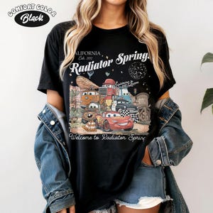 K&ouml;nnte beinhalten: Schwarzes Comfort Colors T-Shirt mit einer Grafik von Charakteren aus dem Film Cars, darunter Lightning McQueen und Mater. Das Design enth&auml;lt den Text "Radiator Springs" und "Welcome to Radiator Spring."