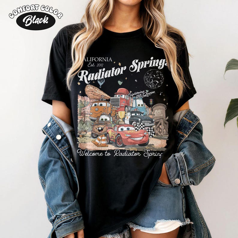 K&ouml;nnte beinhalten: Schwarzes Comfort Colors T-Shirt mit einer Grafik von Charakteren aus dem Film Cars, darunter Lightning McQueen und Mater. Das Design enth&auml;lt den Text "Radiator Springs" und "Welcome to Radiator Spring."