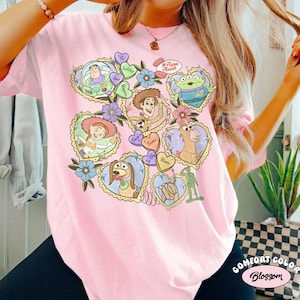 May include: A light pink t-shirt featuring a colorful graphic of characters from the animated film Toy Story. The design includes Buzz Lightyear, Woody, and other characters in heart-shaped frames. The text "Pizza Planet" is also visible.