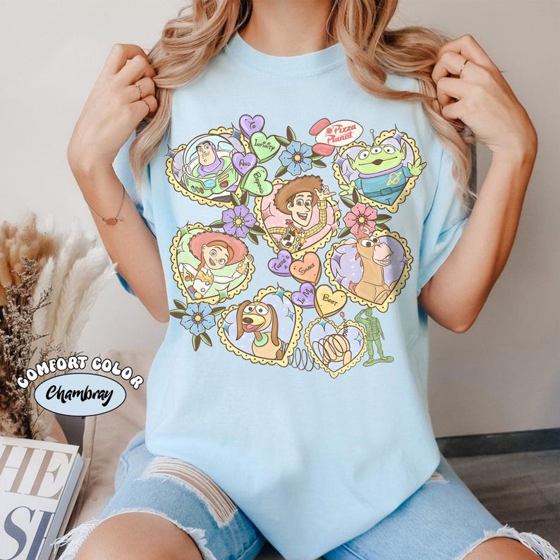 May include: Light blue t-shirt featuring various Toy Story characters in heart-shaped designs. The design includes Buzz Lightyear, Woody, Jessie, Slinky Dog, and others, with floral and heart accents. The shirt has the words "Comfort Color Chambray".