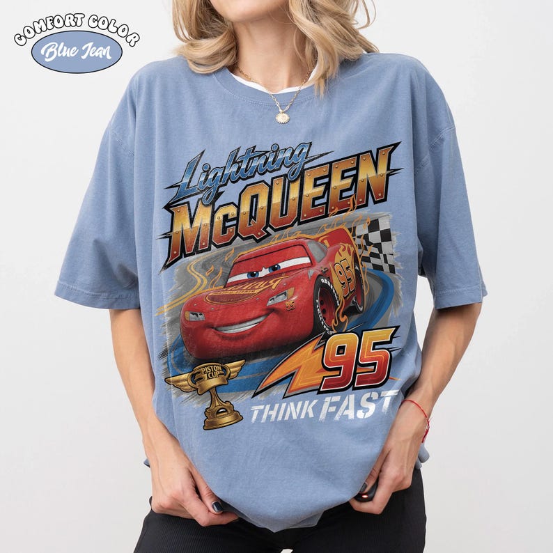 Op de afbeelding: Lichtblauw T-shirt met Lightning McQueen uit de film Cars. De afbeelding bevat de afbeelding van McQueen, het nummer 95 en de zin "THINK FAST". Het shirt heeft ook de woorden "COMFORT COLOR" en "Blue Jean" erop.