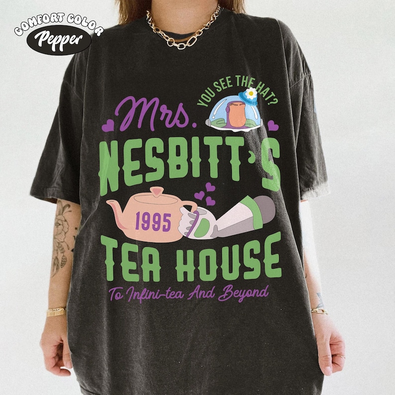 May include: Dark gray Comfort Colors t-shirt with a graphic design. The design features the text "Mrs. Nesbitt's Tea House" in green and purple, along with a teapot, the year 1995, and the phrase "To Infini-tea And Beyond."