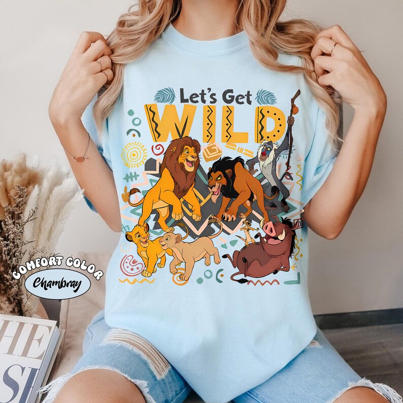 May include: Light blue t-shirt featuring a colorful graphic of characters from The Lion King, including lions, a warthog, and a meerkat. The text "Let's Get WILD" is printed above the characters. The shirt is labeled "Comfort Color Chambray".