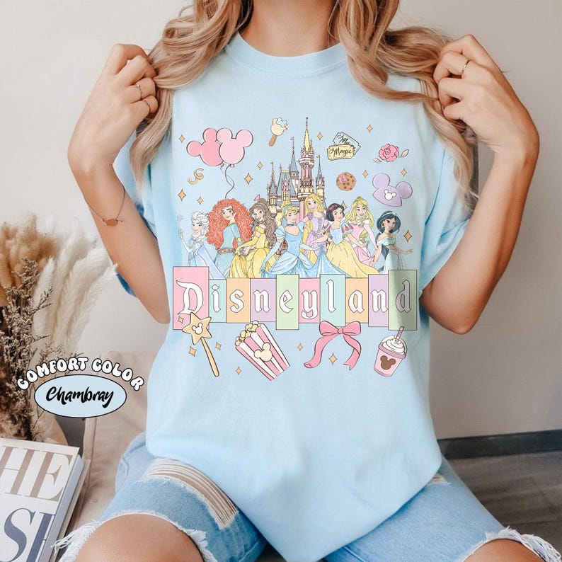 May include: Light blue t-shirt featuring a Disneyland graphic. The graphic includes illustrations of Disney princesses, a castle, and the word "Disneyland" in pastel colors. Additional elements include a wand, popcorn, and a bow.