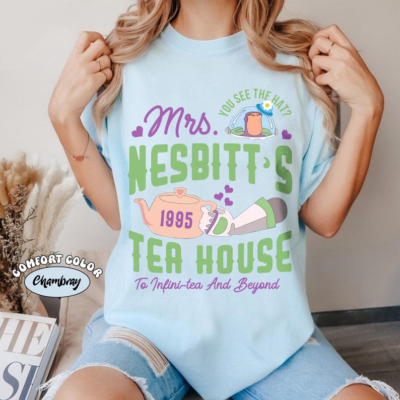 May include: Light blue t-shirt with a graphic design featuring the text "Mrs. Nesbitt's Tea House" and a teapot illustration. The design includes the year 1995 and the phrase "To Infini-tea And Beyond."