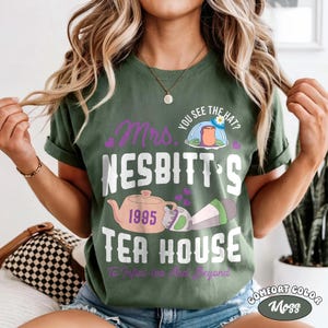 May include: Moss green t-shirt with the text "Mrs. Nesbitt's Tea House 1995" in white and purple. The design includes a teapot, a hat, and the phrase "To Infini-tea And Beyond". The shirt is Comfort Color.