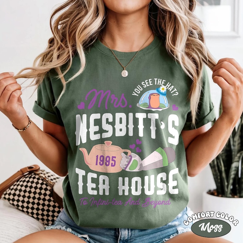 May include: Moss green t-shirt with the text "Mrs. Nesbitt's Tea House 1995" in white and purple. The design includes a teapot, a hat, and the phrase "To Infini-tea And Beyond". The shirt is Comfort Color.