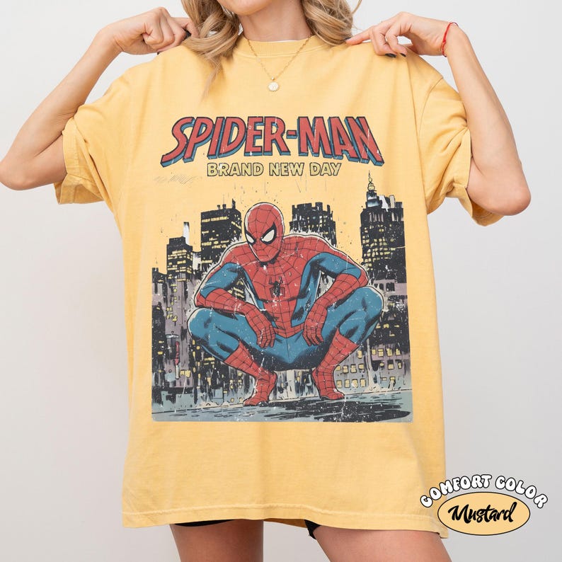 Puede incluir: Camiseta de color mostaza con un gr&aacute;fico de Spider-Man agachado frente a un paisaje urbano. El texto "SPIDER-MAN BRAND NEW DAY" est&aacute; encima de la ilustraci&oacute;n. La camiseta est&aacute; etiquetada como "COMFORT COLOR" y "Mustard".