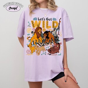 May include: A light purple Comfort Colors t-shirt featuring a graphic of characters from The Lion King. The phrase "Let's Get WILD" is printed above the characters. The design includes Simba, Mufasa, Scar, Timon, Pumbaa, and Zazu.