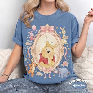 May include: Blue jean Comfort Colors t-shirt with a framed illustration of Winnie the Pooh and friends. The design includes Piglet, Tigger, Eeyore, and Rabbit, with pink bows, flowers, and bees. The shirt has rolled-up sleeves.